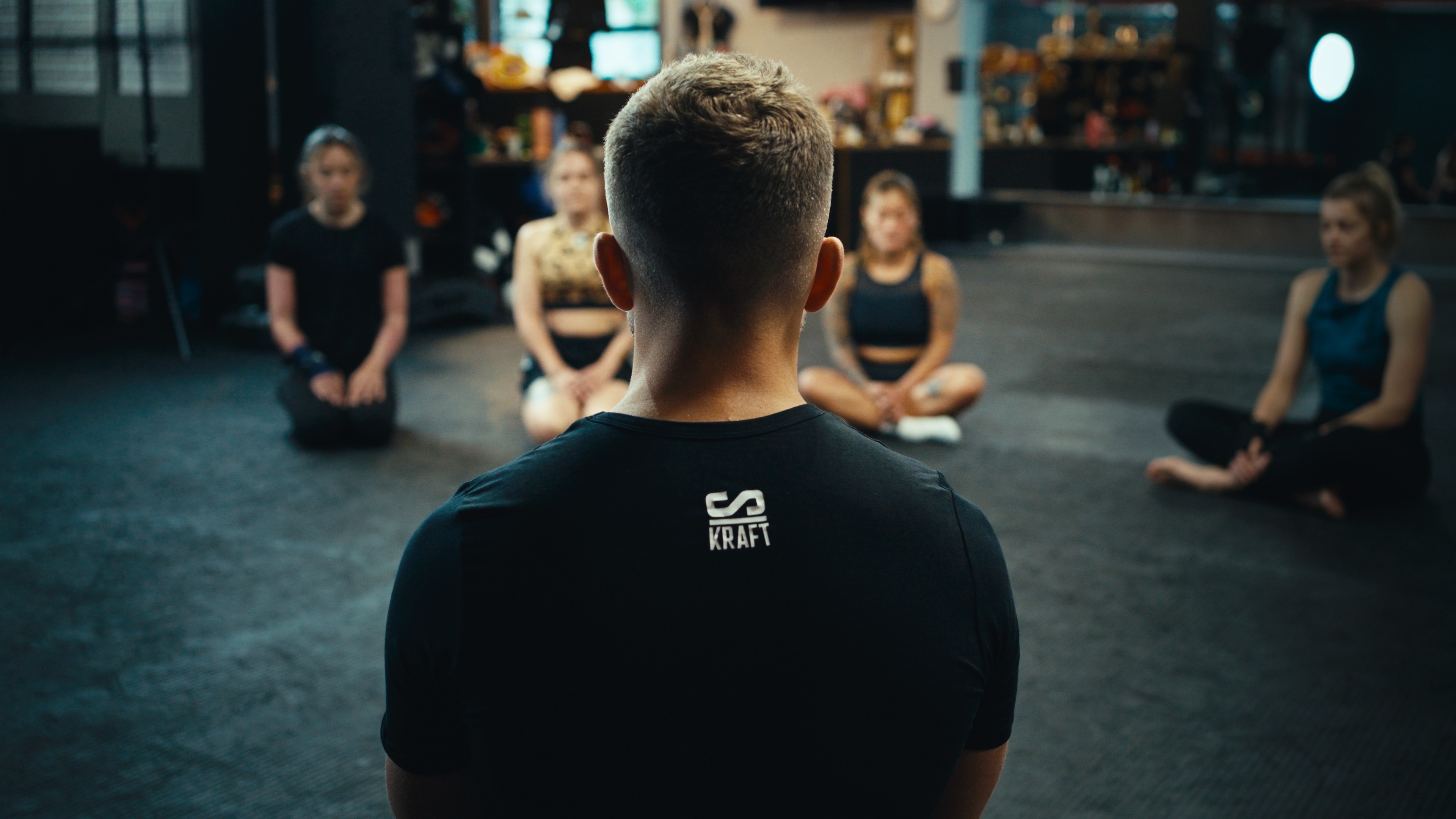 Coach im Schlagkraft-Shirt vor einer im Kreis sitzenden Teamgruppe im Boxgym